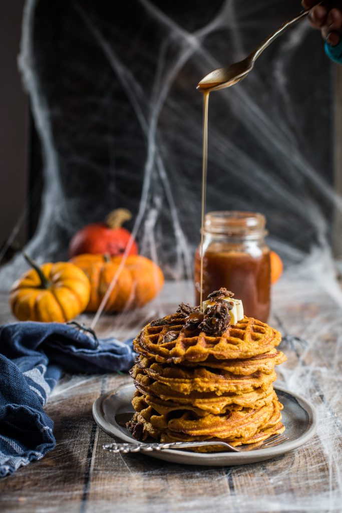 Pouring homemade pumpkin pie spice syrup over pumpkin waffles with caramelized pumpkin seeds